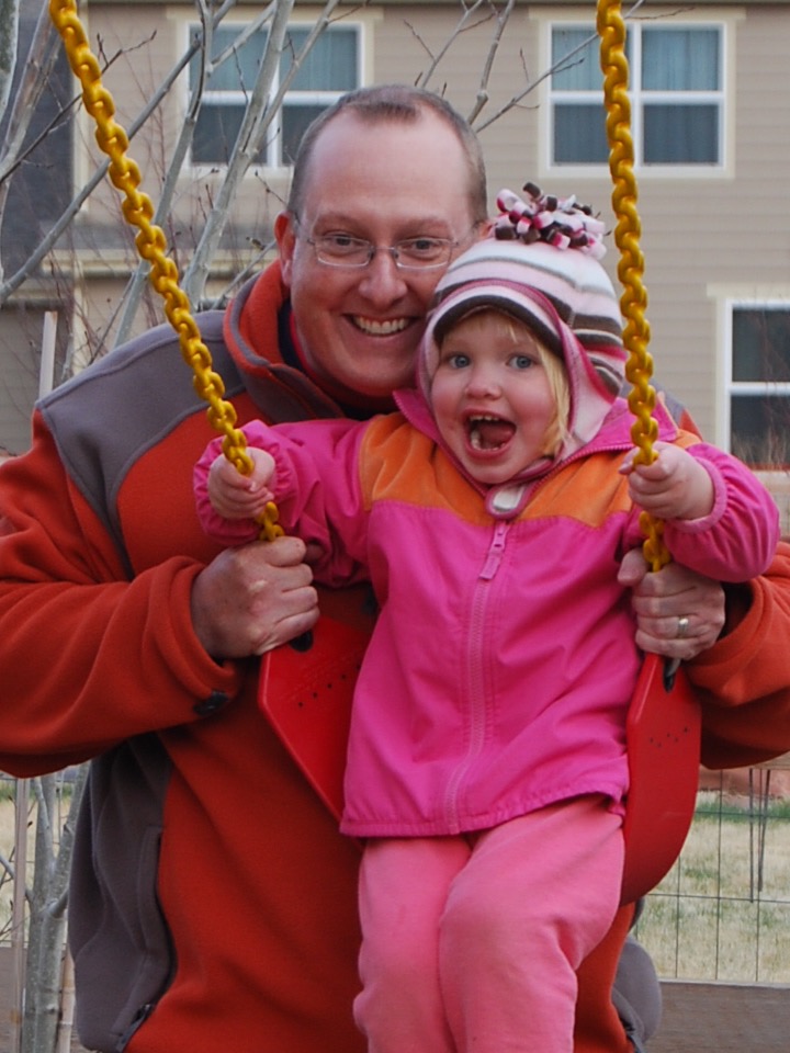 Emma as a toddler, grinning on a backyard swing while her dad holds the chains behind her.